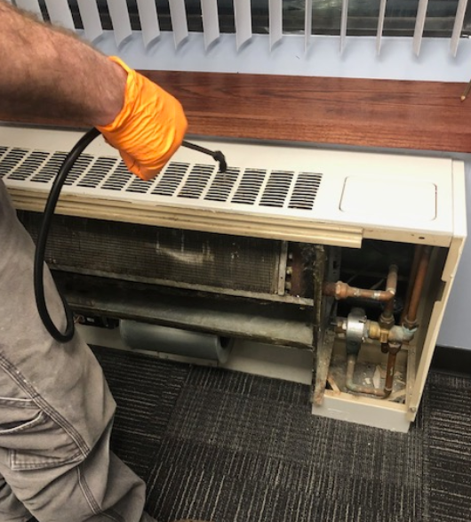 A maintenance worker in orange gloves is using a cleaning tool to service a dirty fan coil unit.