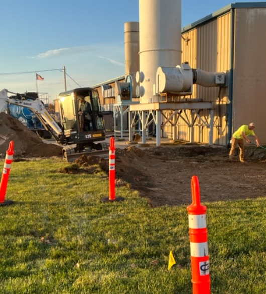 An image of two construction workers and a small excavator working outside a commercial building.